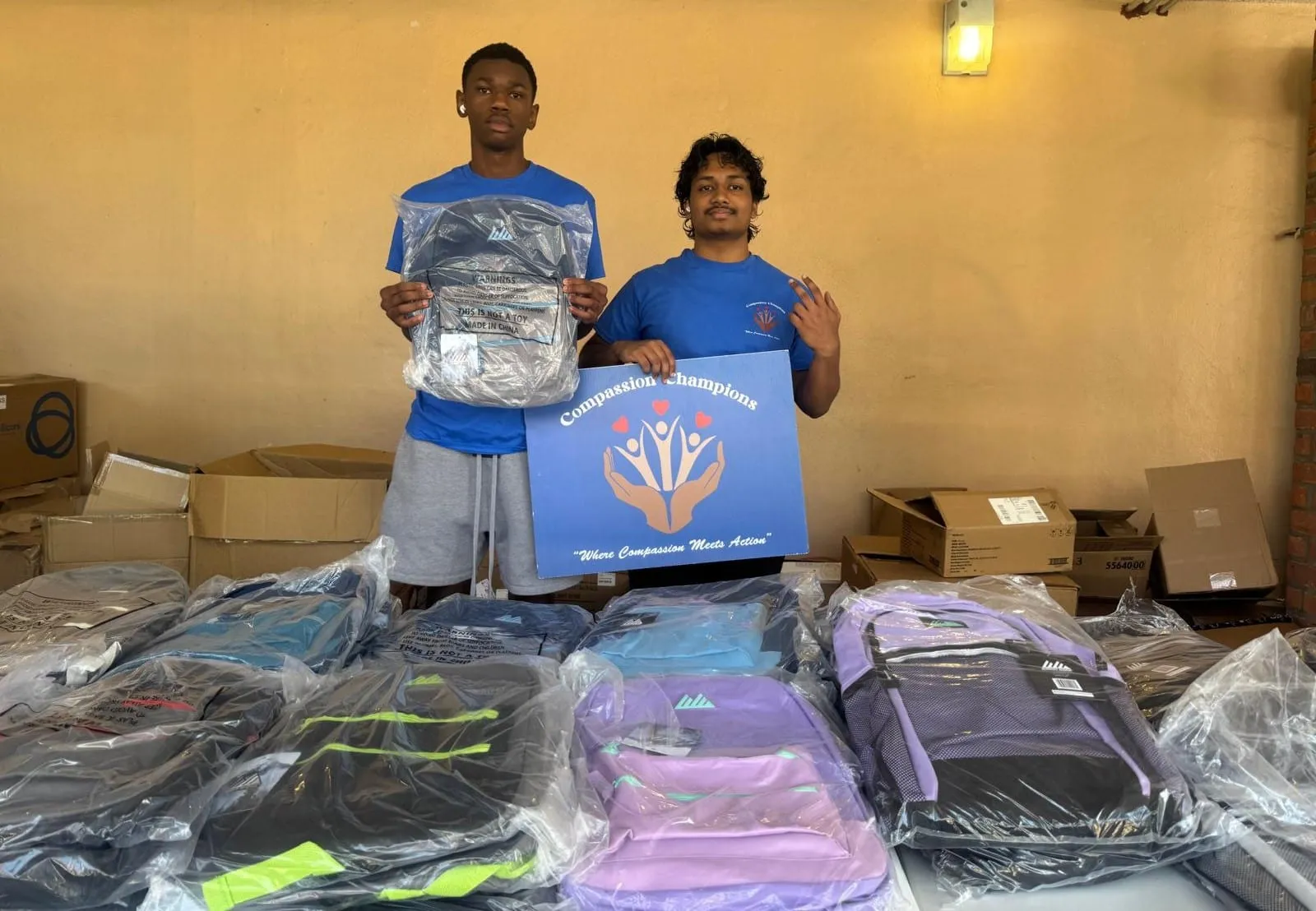 Two volunteers with the Compassion Champions sign behind rows of backpacks